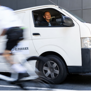 Smiling driver in an EZCourier van waits at the wheel as a cyclist passes by in the foreground.